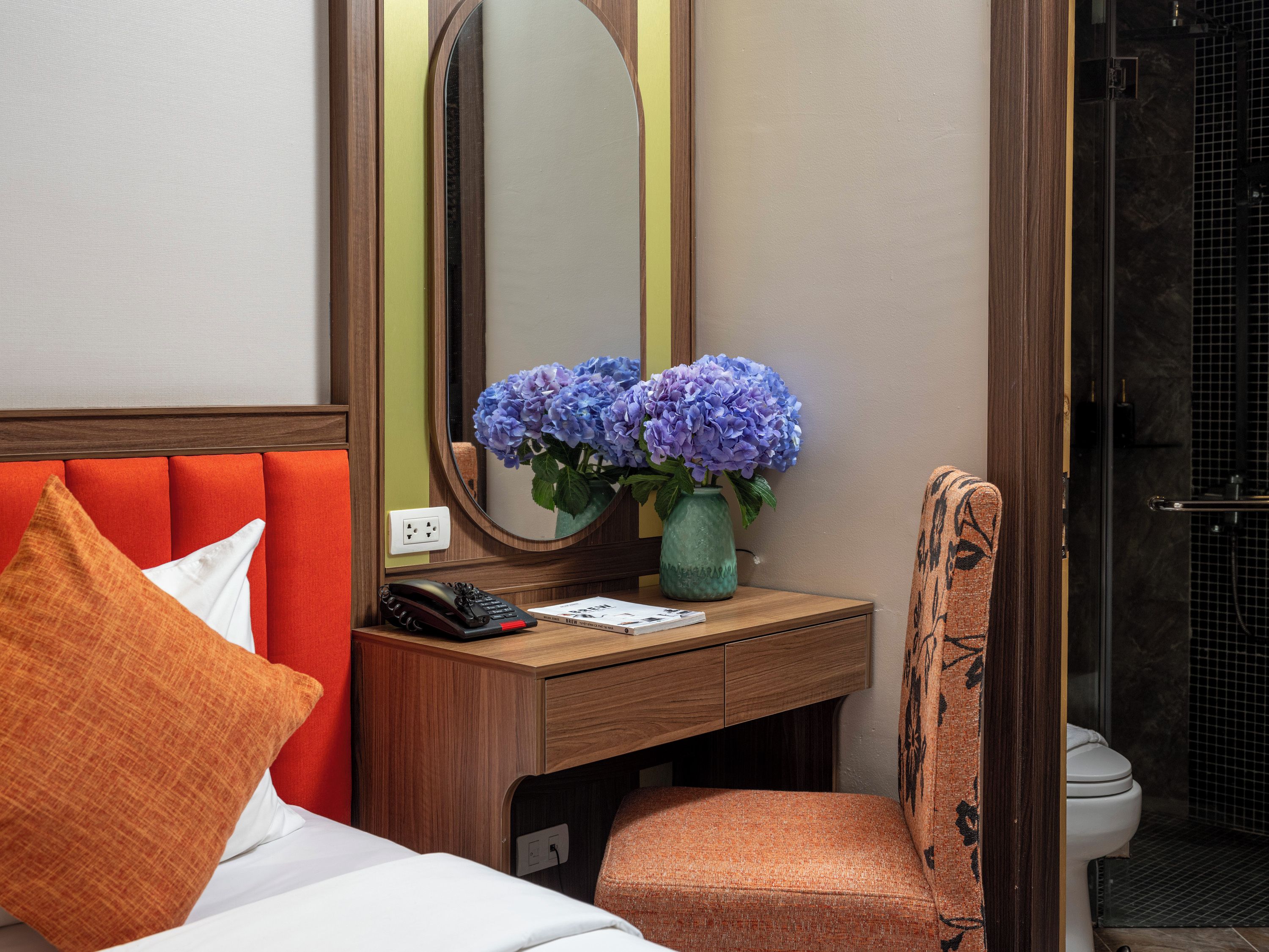 A hotel room with a bed, a wooden desk with a mirror, a vase with flowers, a telephone, and a bathroom visible through an open door.