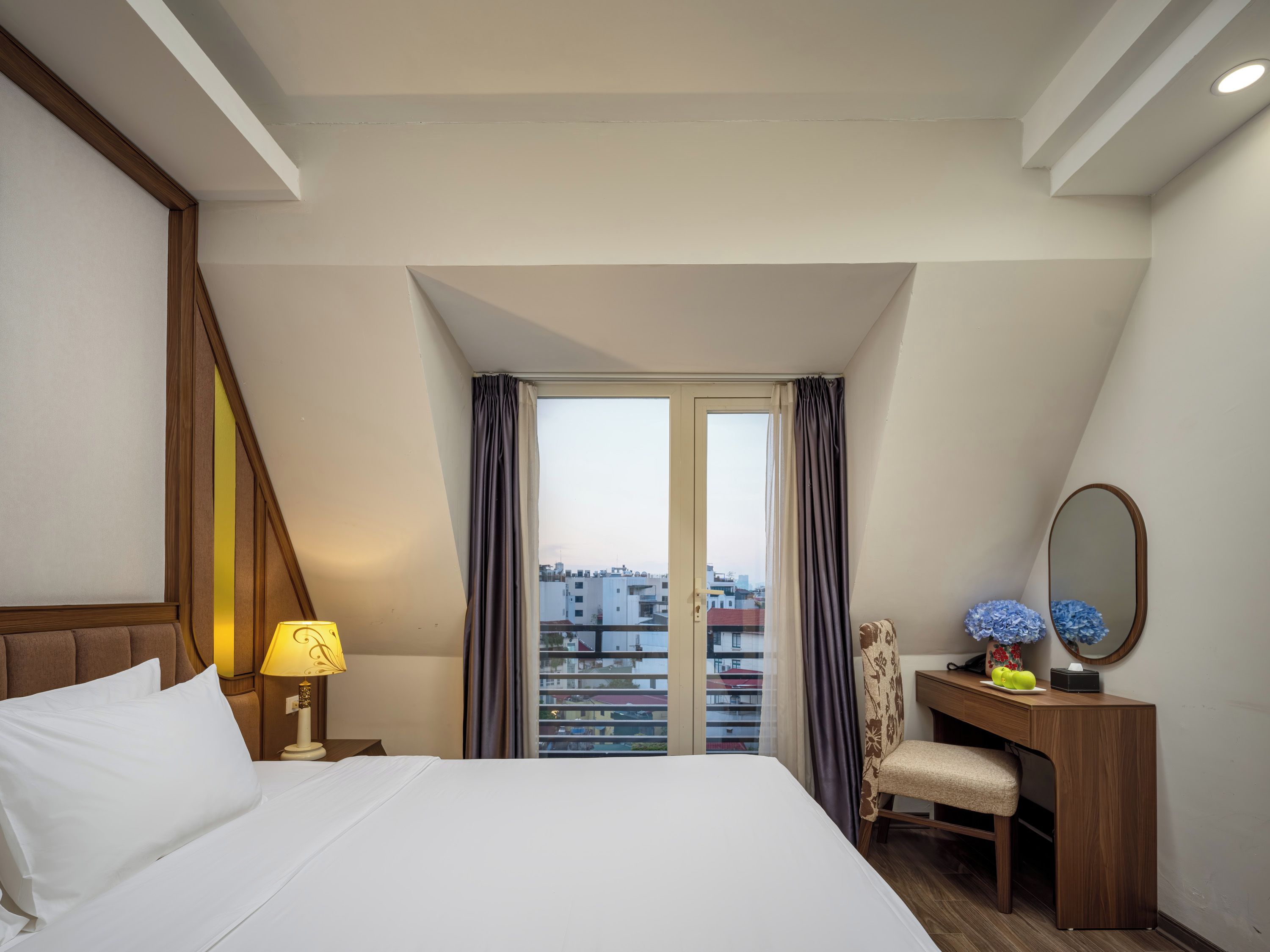 Habitación de hotel con una cama, un escritorio, una silla, un espejo y vistas a edificios a través de una ventana.