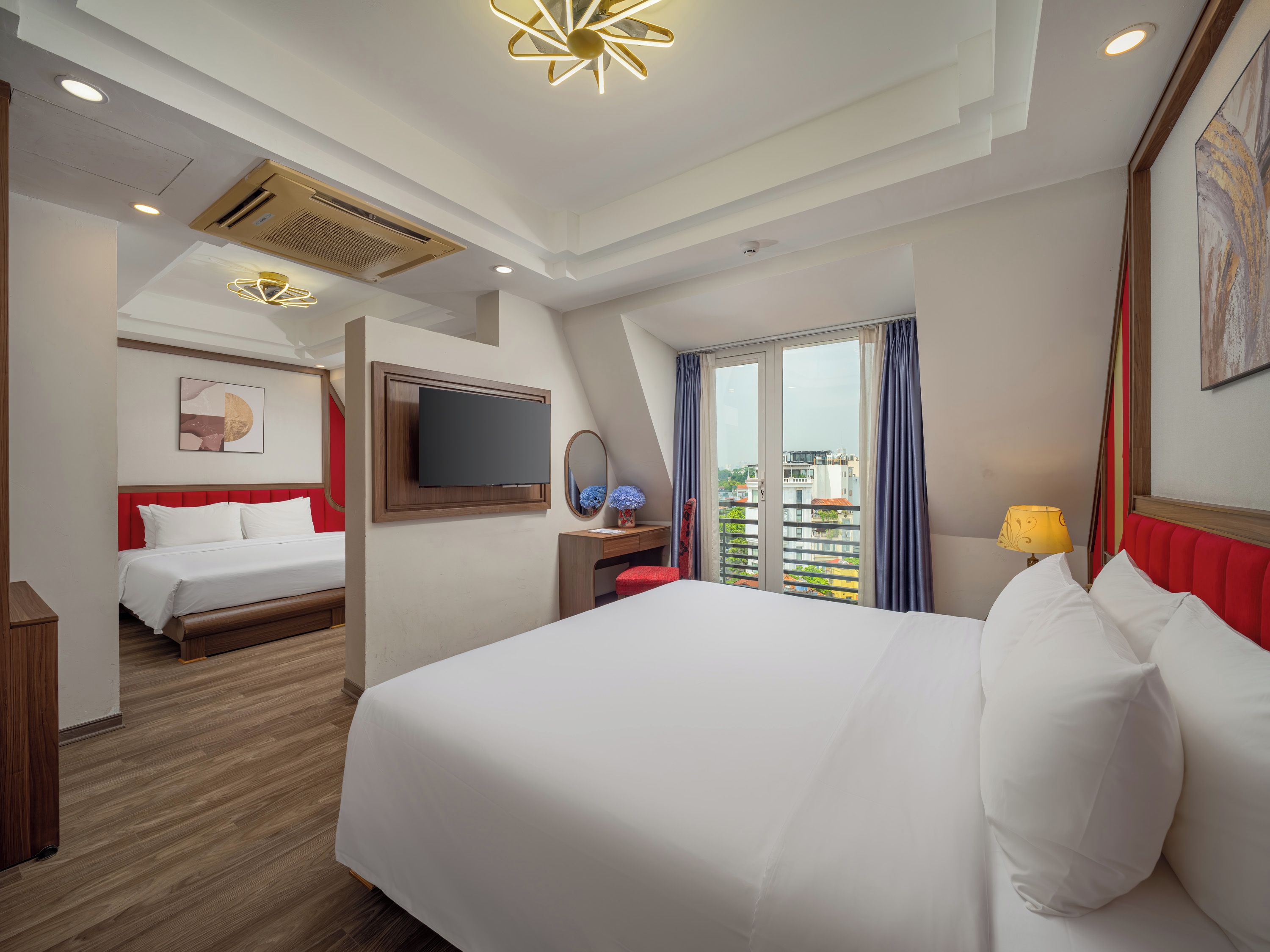 A hotel room with a large bed, a TV mounted on the wall, a desk with a lamp, and a view of the city through the window.