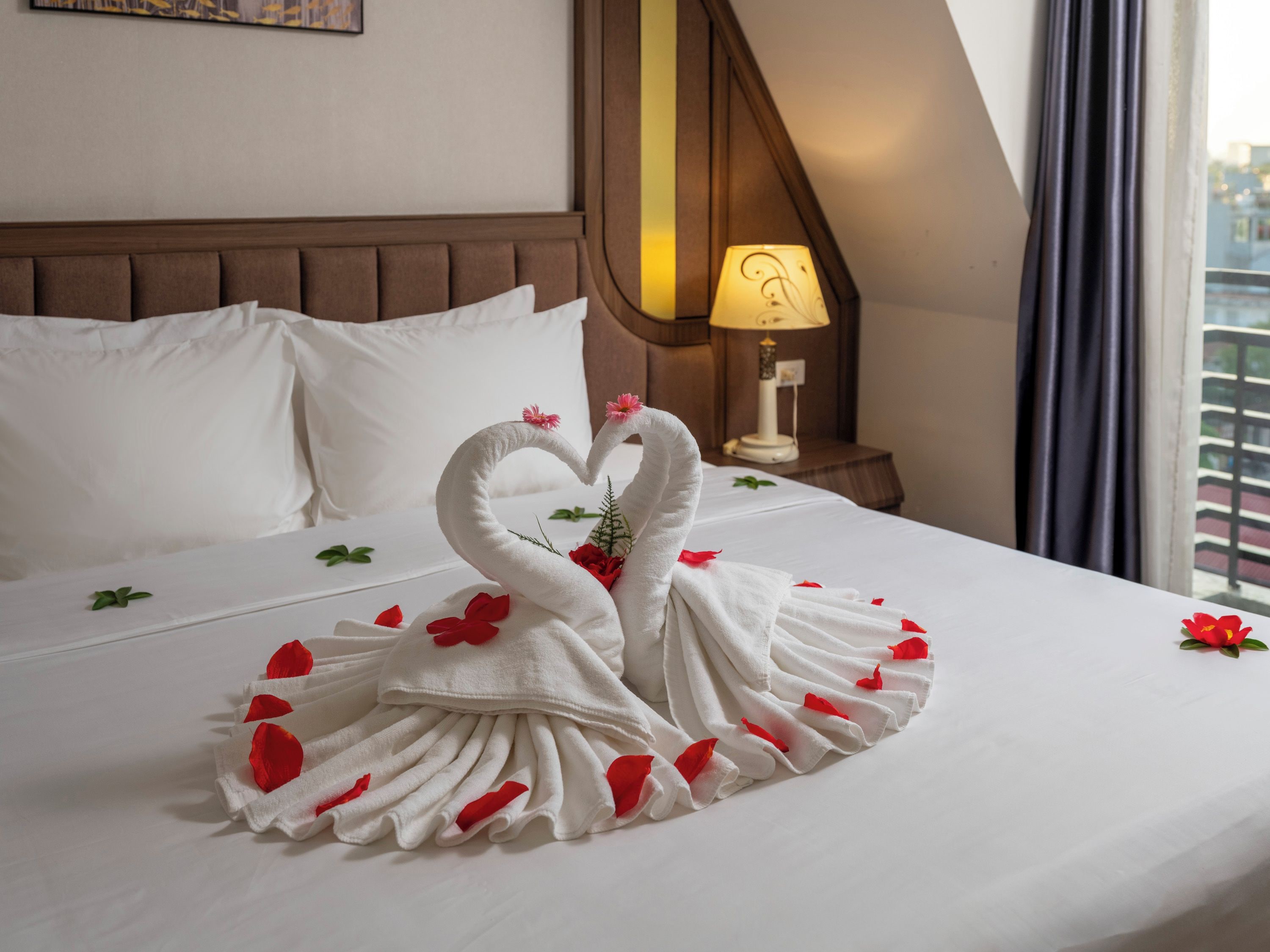 A hotel room with a bed, pillows, a nightstand with a lamp, and a window with curtains.