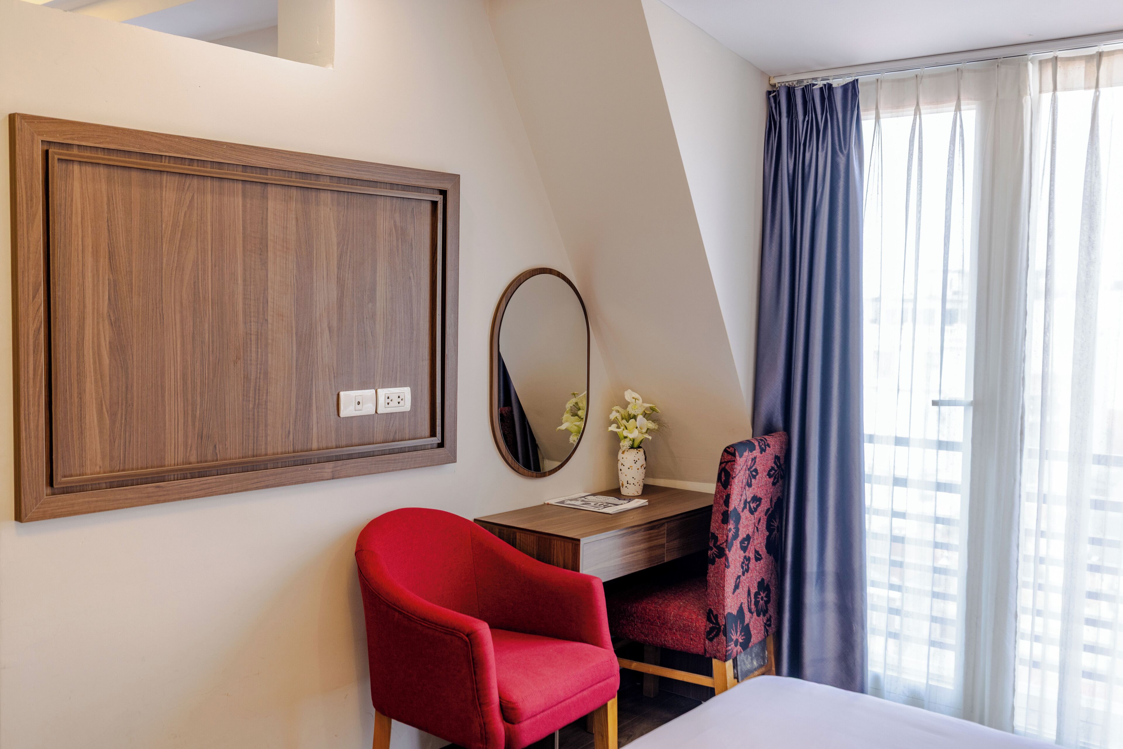A hotel room with a wooden wardrobe, a desk with a mirror, a red chair, a bed, and a window with curtains.