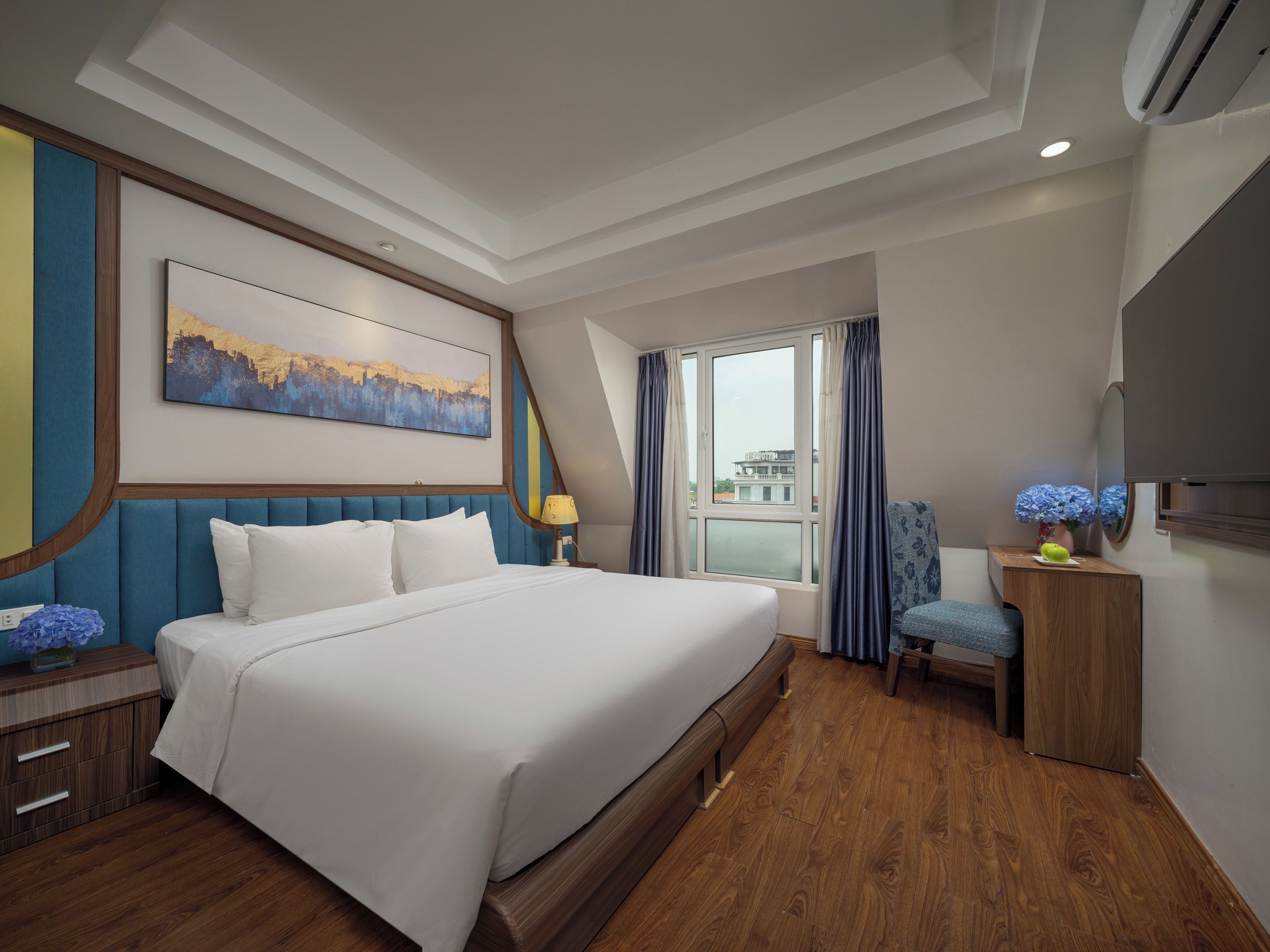 Quarto de hotel com uma cama grande, uma escrivaninha, uma cadeira, uma janela com cortinas, um quadro na parede e um aparelho de ar condicionado.
