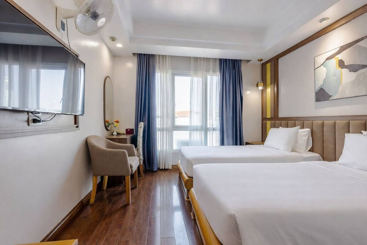 Une chambre d’hôtel avec deux lits, un bureau avec une chaise, un téléviseur fixé au mur et une grande fenêtre avec des rideaux.