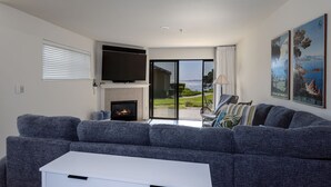 Ein modernes Wohnzimmer mit grauem Sofa, einem Flachbildfernseher, einem Kamin und Meerblick durch große Glasfenster.