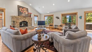 A spacious living room with a stone fireplace, grey sofas, a flat-screen TV, and large windows.
