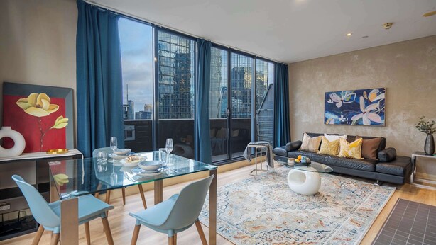 Een moderne woonkamer met een glazen eettafel, blauwe stoelen, een bank met kussens, een salontafel en een groot raam met gordijnen.
