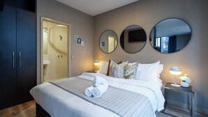 Een opgemaakt bed met opgerolde handdoeken, een bedlampje en een nachtkastje met een bloevas.