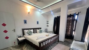 A hotel room with a bed, bedside tables, a nightstand, a small table, a chair, a window with curtains, and a wall-mounted light fixture. - Thar View Hotel (Jaisalmer)