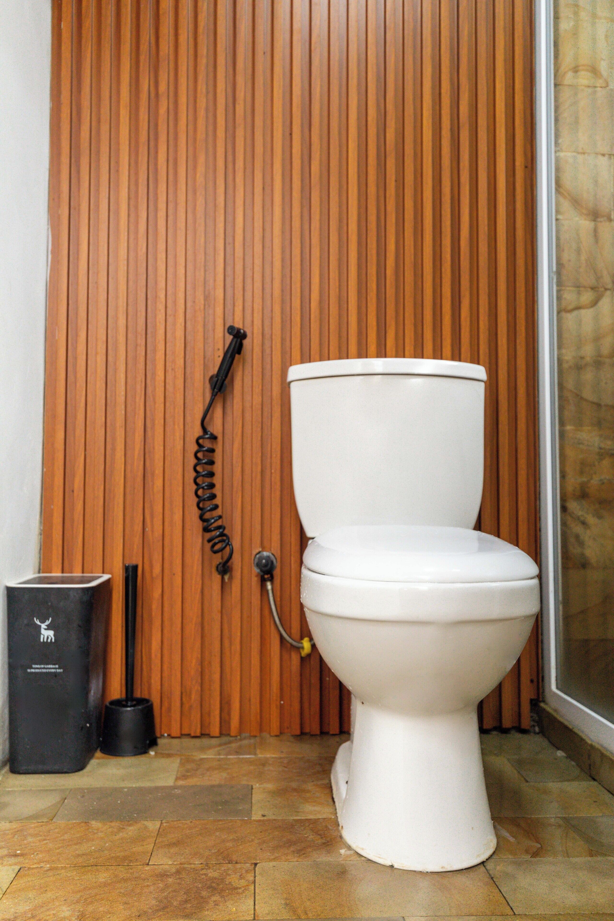 Des toilettes blanches, avec un mur en lambris de bois derrière, une poubelle noire et un sol recouvert de carreaux à motifs.