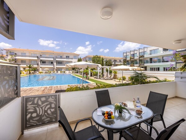 A balcony with a pool view, outdoor seating, and a table with fruit and drinks. - 2bedroom apartment in kapparis Mythical AL04 (Kapparis)