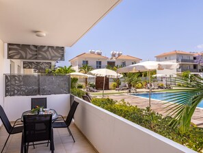 A balcony with a table and chairs, overlooking a swimming pool and adjacent buildings. - 2bedroom apartment in kapparis Mythical AL04 (Kapparis)