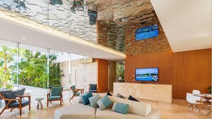 A modern hotel lobby with a large sectional sofa, wooden paneling, a TV screen, and a glass wall overlooking a lush garden. - Alba Puerto Cancún, Modern Lifestyle with Rooftop Near the Marina (Puerto Cancun)