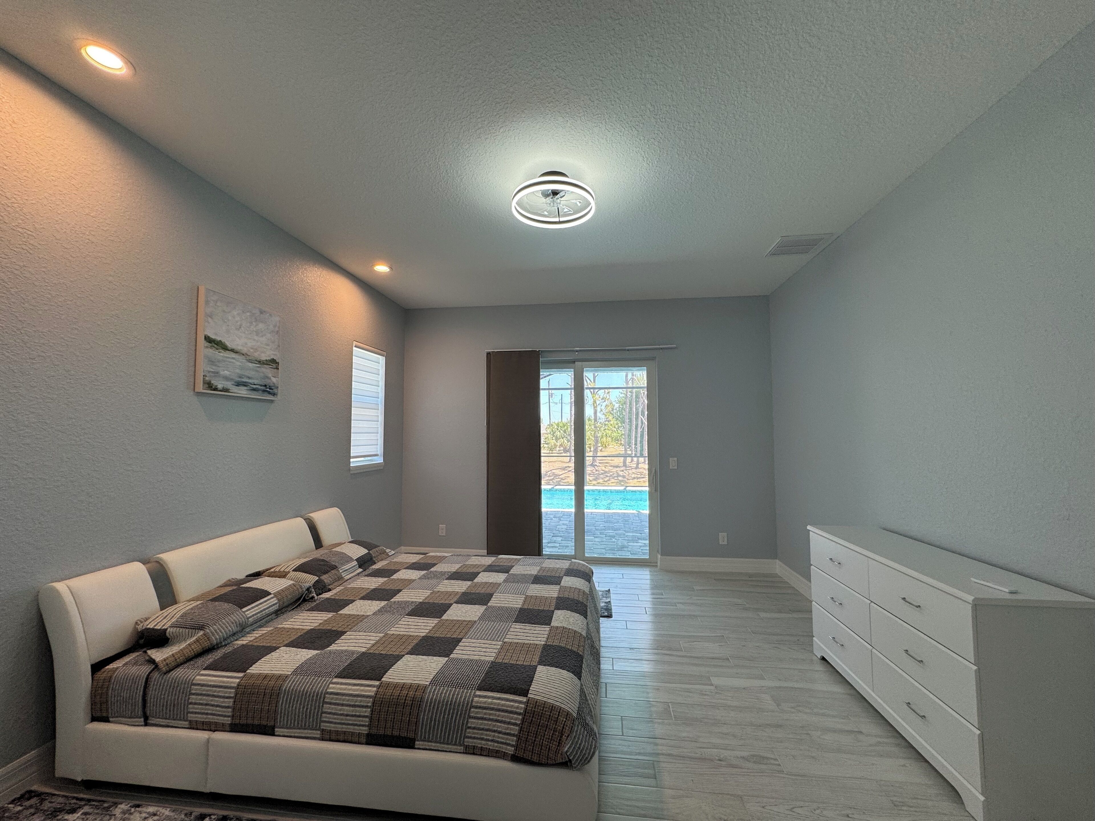 A bedroom with a bed, a dresser, a painting, and a view of a swimming pool through a glass door.