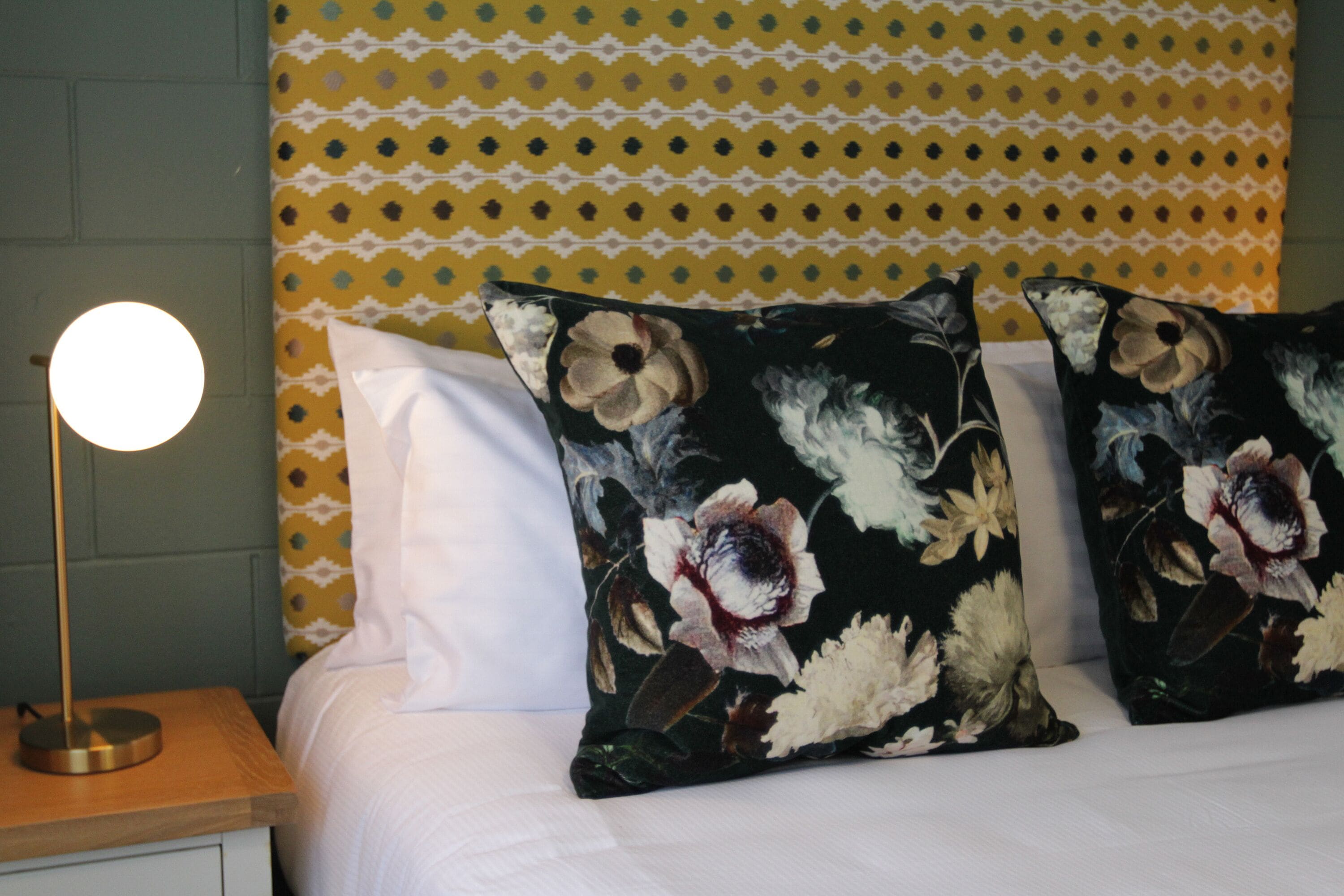 A bed with white bedding, two floral pillows, a round table lamp, and a patterned wall behind.