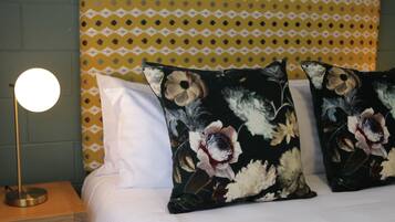 A bed with white bedding, two floral pillows, a round table lamp, and a patterned wall behind.