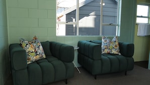 Deux fauteuils verts ornés de coussins à motifs floraux, une fenêtre donnant sur un immeuble et un porte-serviettes avec une serviette.