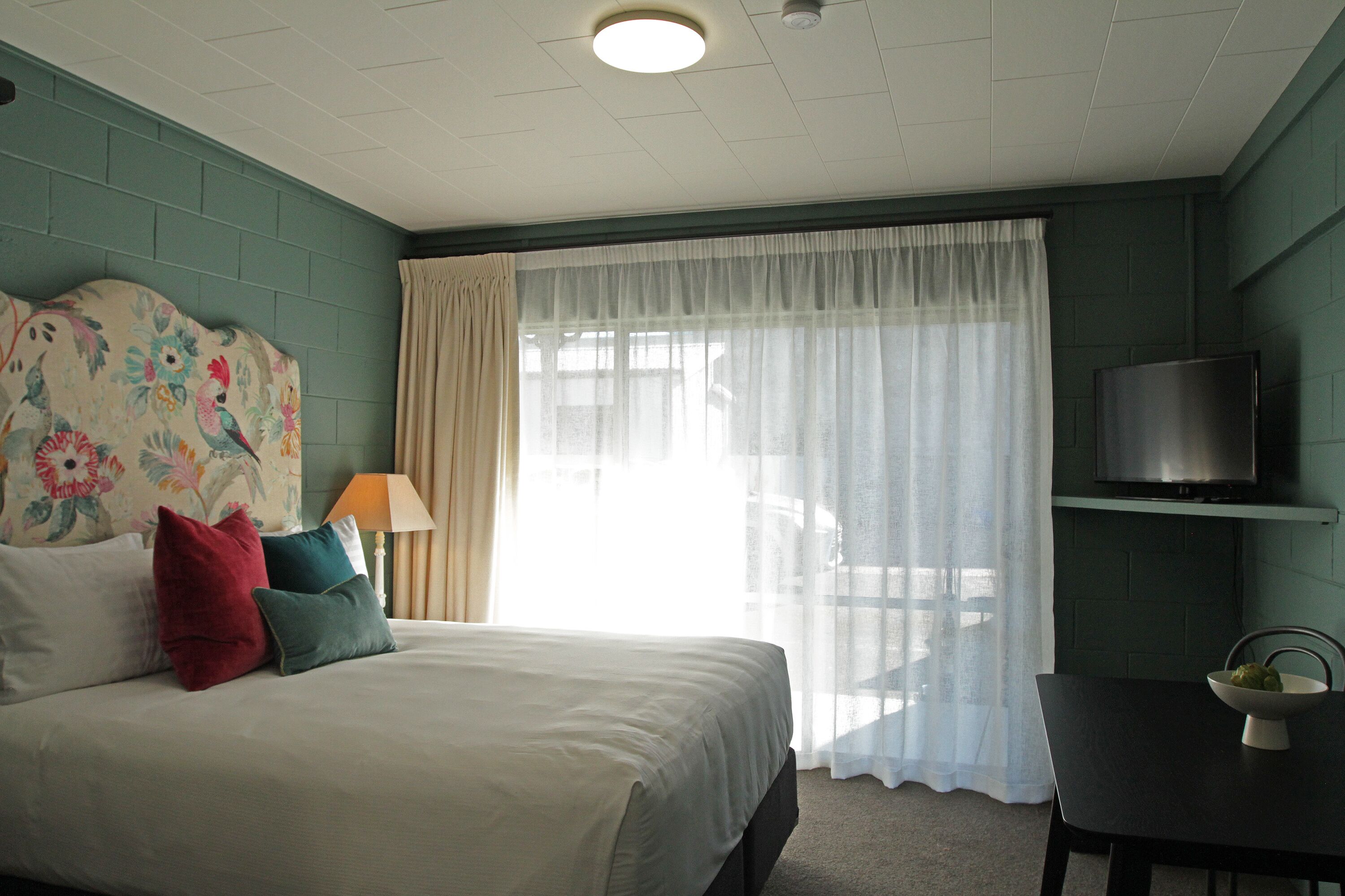 A hotel room with a bed, a television on a stand, a table with a bowl, and a window with curtains.