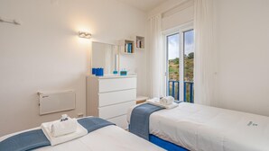 Ett hotellrum med två sängar, en vit byrå, en spegel, ett fönster med gardiner och utsikt över omgivningarna.