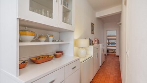 Eine schmale Küche mit weißen Schränken, gefliestem Boden und einer Arbeitsplatte mit Geschirr und einer Lampe.