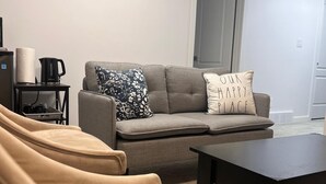 Una sala de estar con un sofá gris, una mesa de centro negra y un sillón beige. Hay un cuadro en la pared y una puerta al fondo.