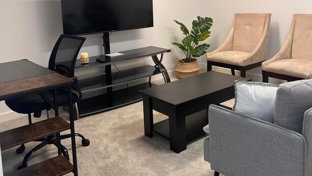 Un espacio de oficina moderno con un televisor de pantalla plana grande, un escritorio con una silla, una mesa de centro y dos sillones. Hay una planta pequeña en el escritorio y otra en el suelo.