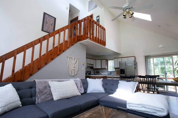 A spacious living area with a grey sectional sofa, wooden staircase, and a kitchen in the background.