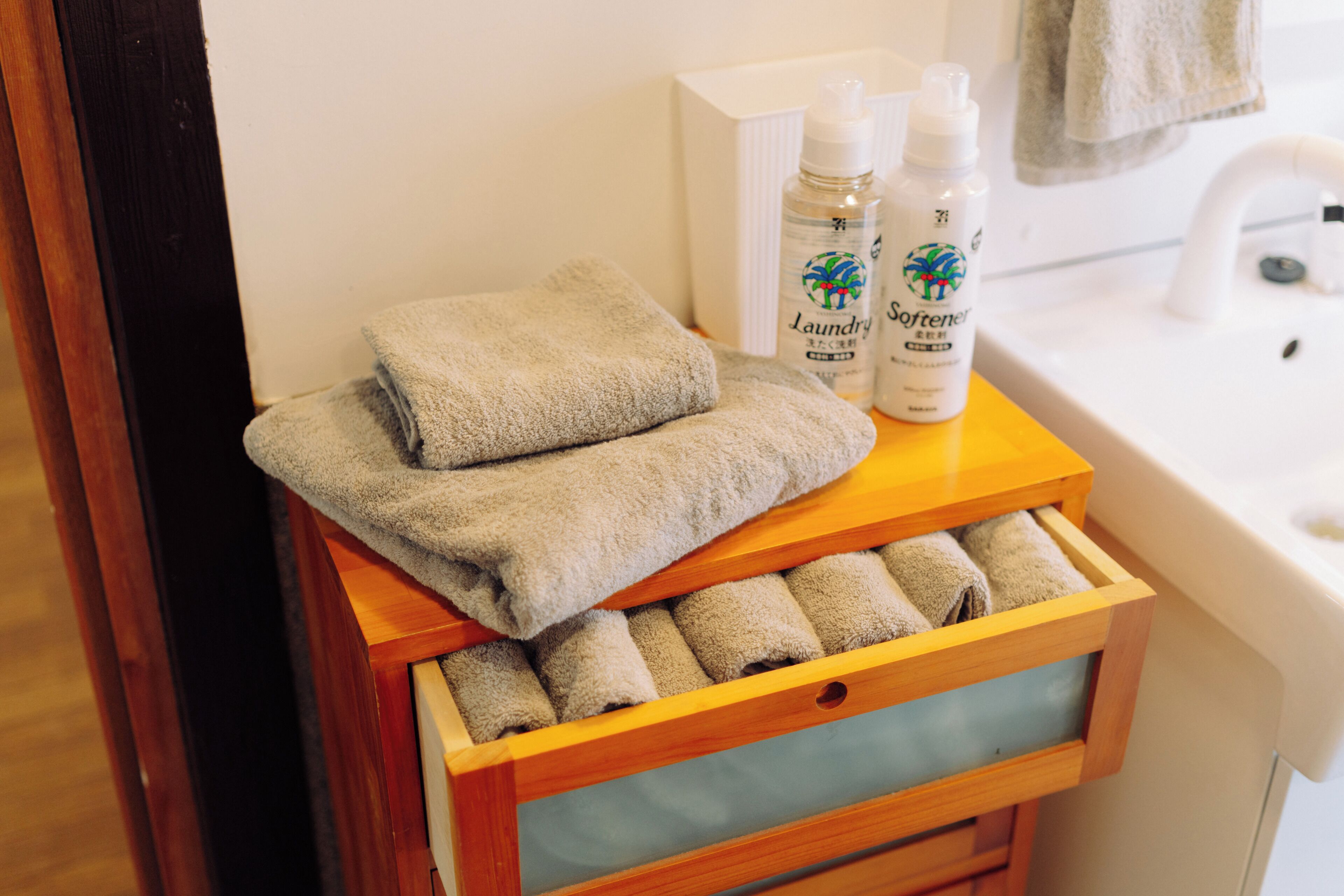 Un tiroir en bois contenant des serviettes soigneusement pliées et deux bouteilles d’adoucissant à linge, posées sur le comptoir d’une salle de bain.