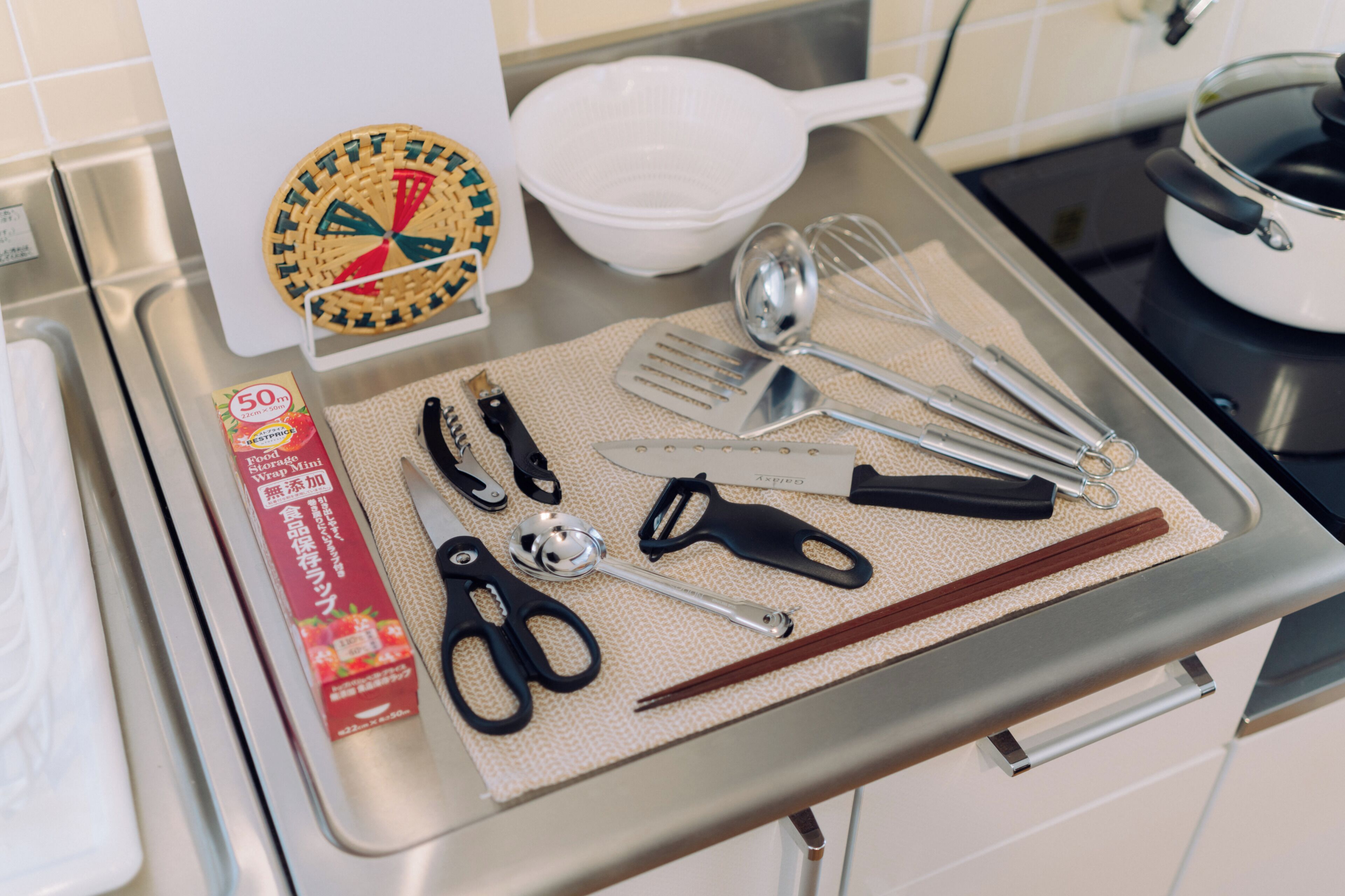 A kitchen counter with various utensils, a white bowl, a pot on the stove, and a box of food items.