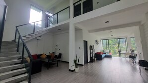 Amplia sala de estar con escalera, un conjunto de sofás, una mesa de comedor y ventanas grandes que ofrecen vistas a zonas verdes.