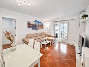 Ein modernes Wohnzimmer mit einem beigefarbenen Sofa, einem Esstisch und einem Fernseher. Ein großes Fenster bietet Blick ins Freie.