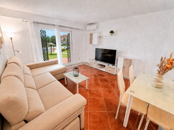 A modern living room with a beige sofa, a TV, a small table, and a dining area with chairs. - Alcaraván T2, ground floor (Ayamonte)