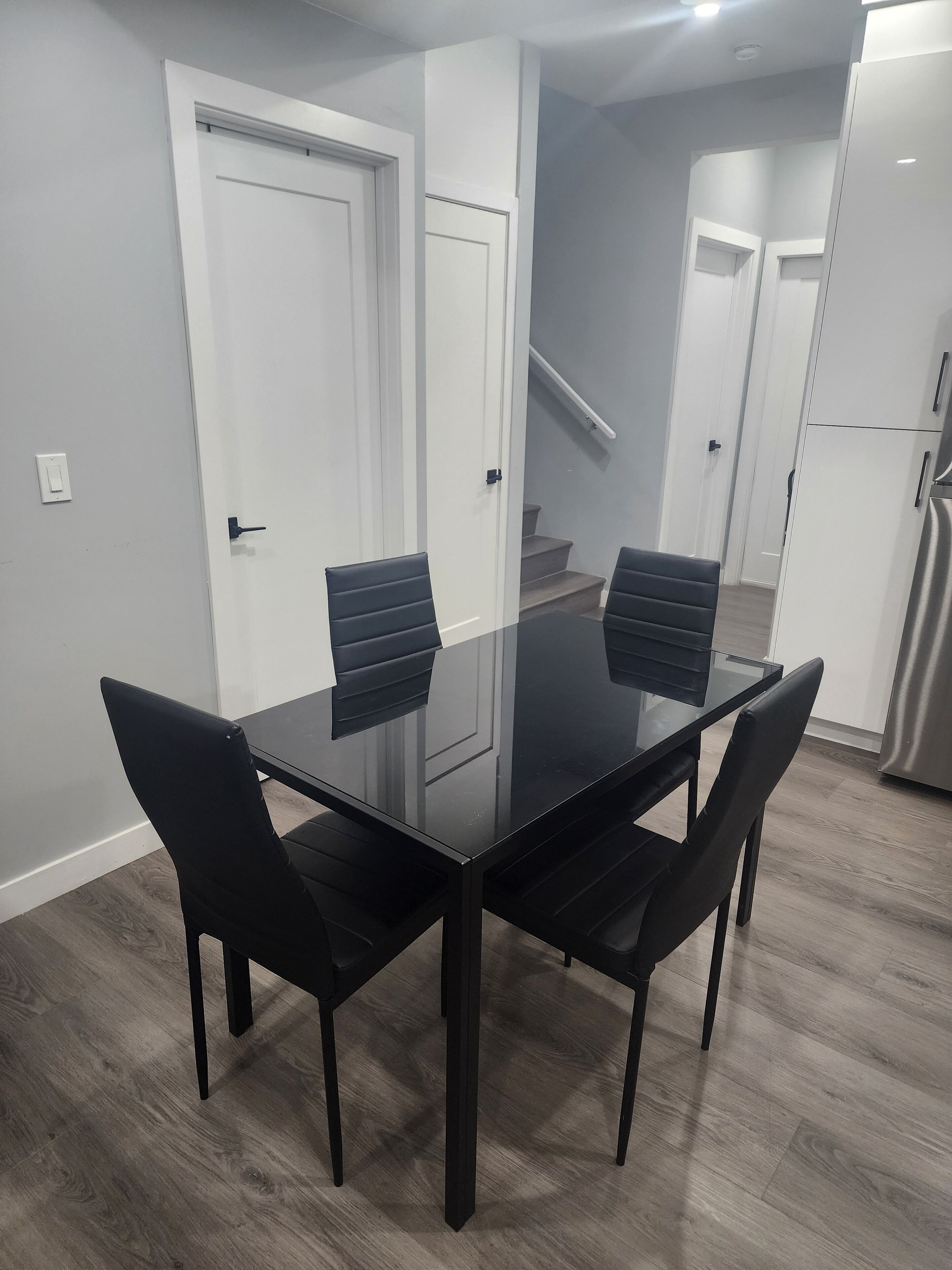 A modern dining area with a glass-top table and black chairs, a white door, and a kitchen with a stainless steel refrigerator.