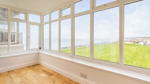 A room with large windows, wooden flooring, and a view of a green field and buildings.