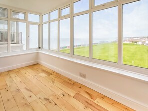 A room with large windows, wooden flooring, and a view of a green field and buildings.
