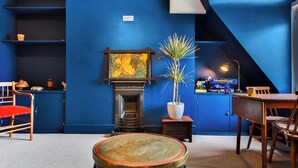 A room with a blue wall, a fireplace, a table, chairs, a small plant, and a shelf with items.