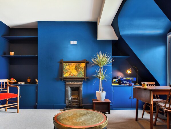 Une pièce avec un mur bleu, une cheminée, une table, des chaises, une petite plante et une étagère avec divers objets.