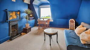 A room with a fireplace, a dining table with chairs, a round coffee table, a sofa with patterned cushions, and a view through a window.
