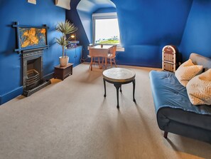 A room with a fireplace, a dining table with chairs, a round coffee table, a sofa with patterned cushions, and a view through a window.