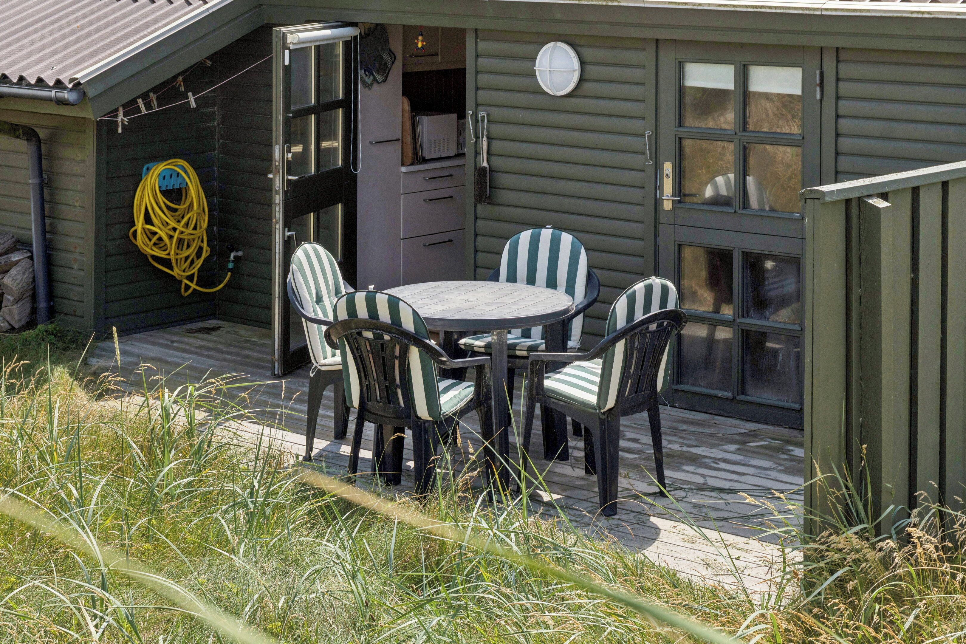 Een houten terras met een ronde tafel en stoelen, een deur naar een garage en een opgerolde tuinslang aan de muur.