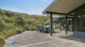 A wooden deck with a table and chairs, a covered area with a grill, and a view of a grassy hill. - Summerhouse in peaceful surroundings (Pandrup)