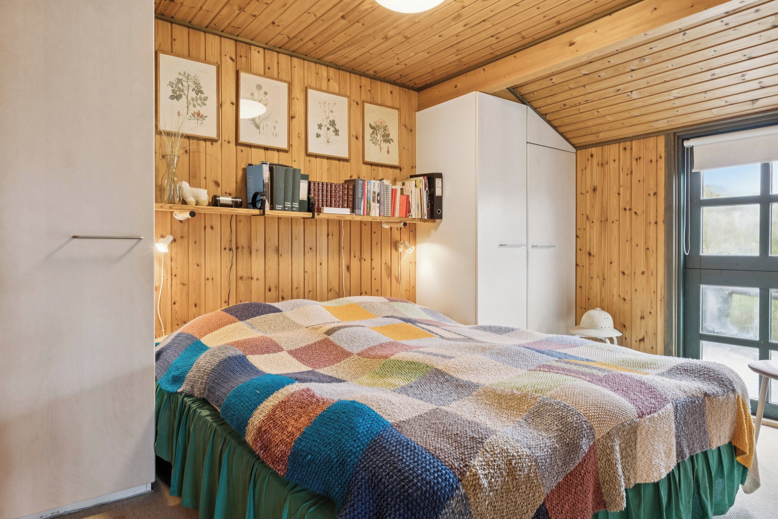 A bedroom with a wooden ceiling, a quilted bedspread, a bookshelf with books, and a window with a view.