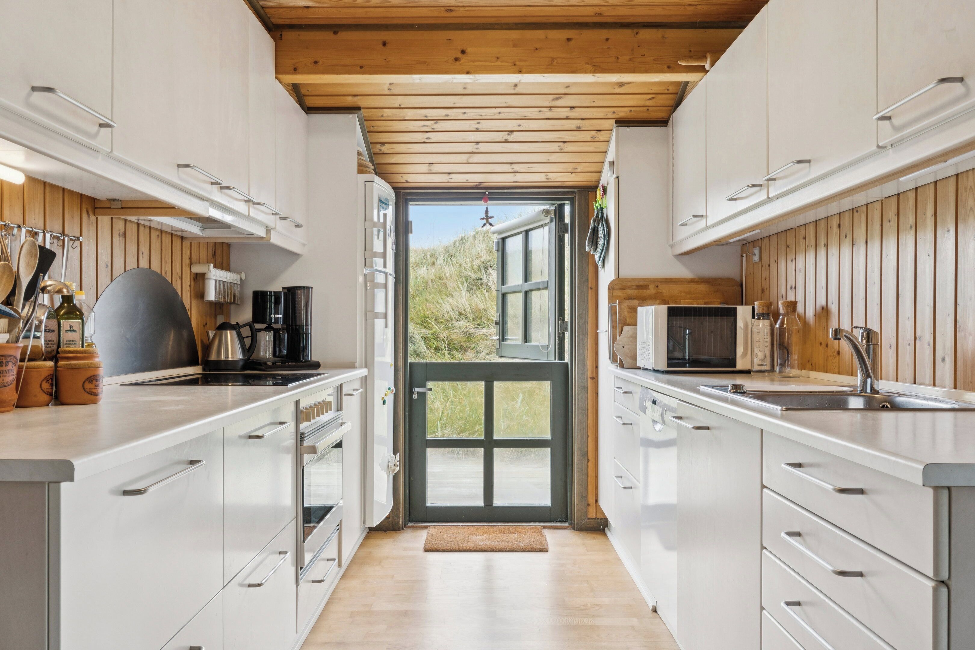 Uma cozinha moderna com armários brancos, teto de madeira e vista através de uma porta de vidro.