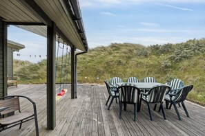 En träveranda med ett bord och stolar, en gunga och utsikt över stranden och sanddynerna.