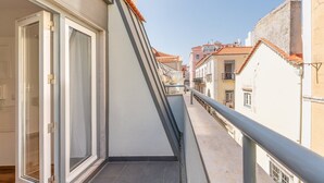 Un balcón estrecho con barandilla metálica, con vistas a una calle con edificios tradicionales y cielo despejado.