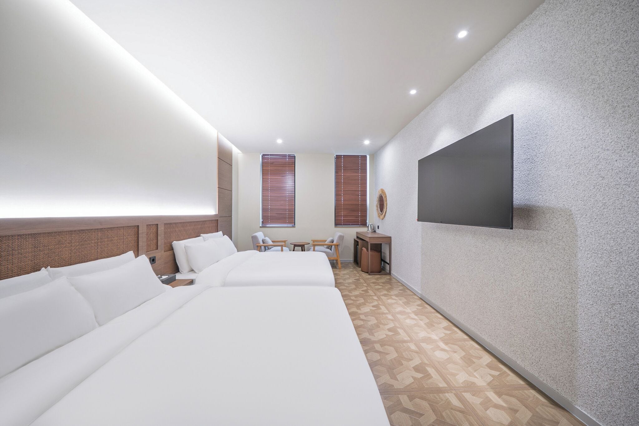 A modern hotel room with a large bed, a desk, two chairs, a flat-screen TV mounted on the wall, and a patterned floor.