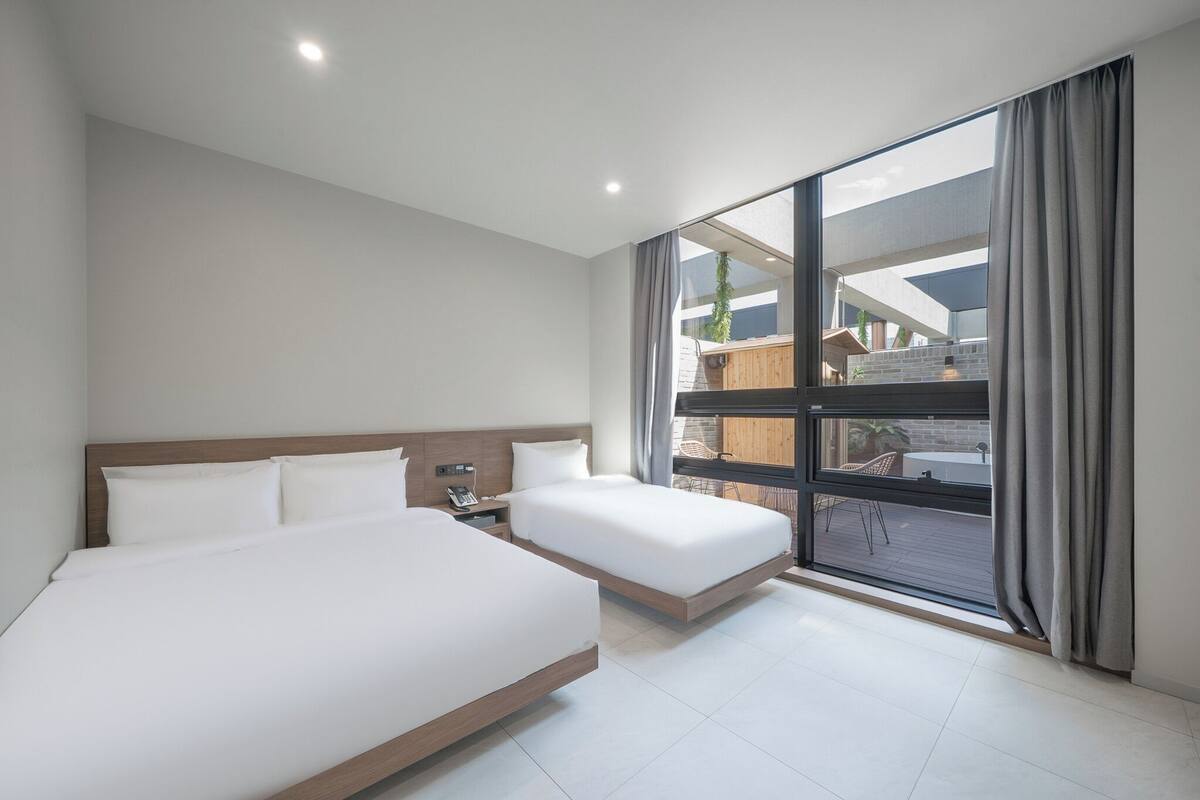 A modern hotel room with two beds, a large window, and a view of the outdoor area.