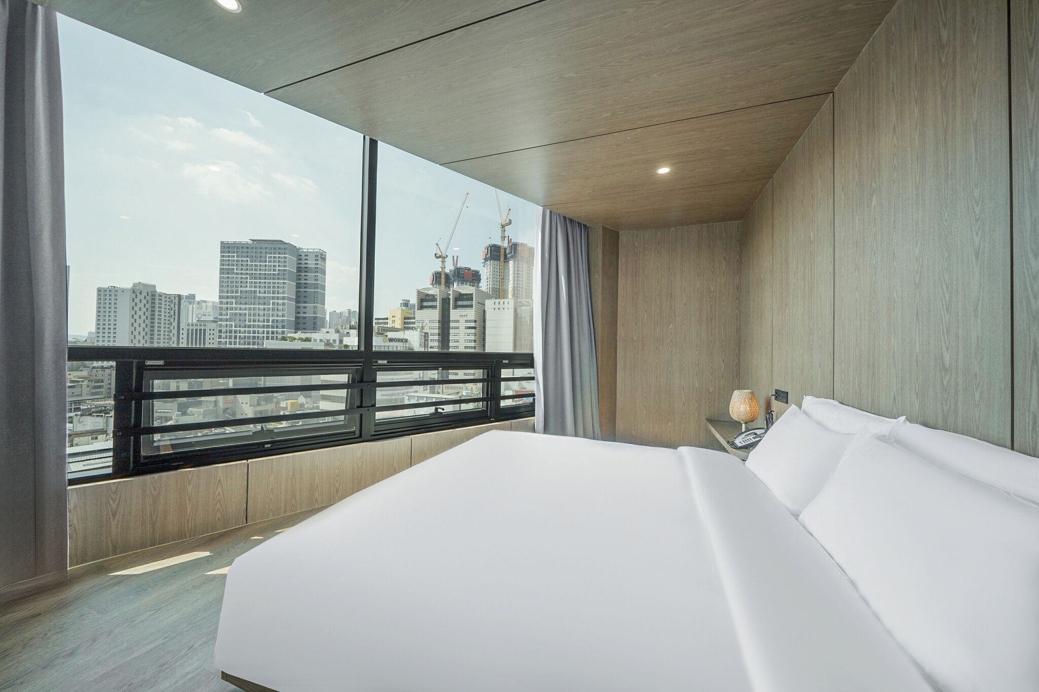 A modern hotel room with a large bed, a nightstand with a lamp, and a view of the city skyline through large windows.