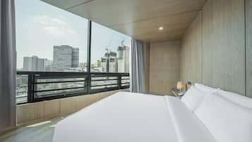 A modern hotel room with a large bed, a nightstand with a lamp, and a view of the city skyline through large windows.