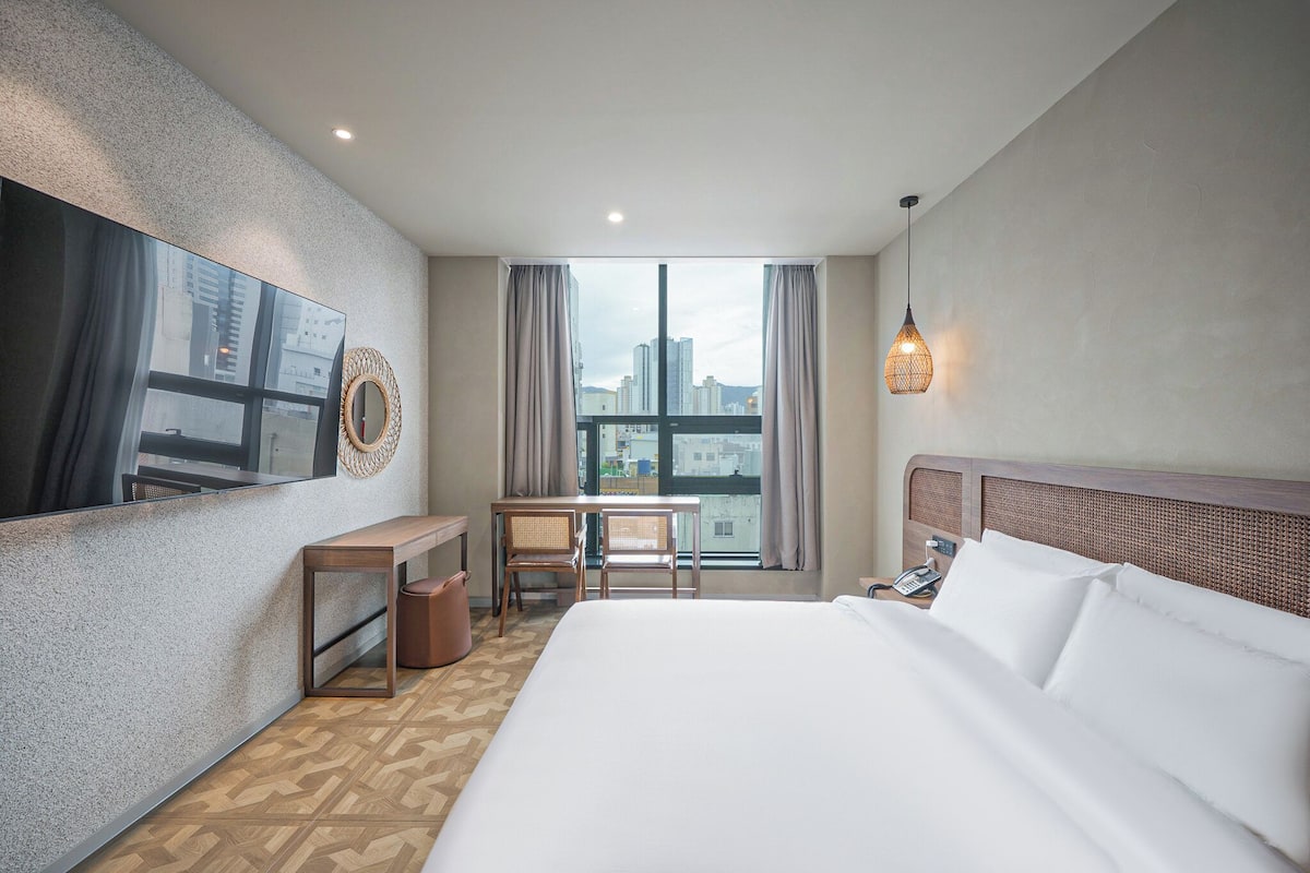 A hotel room with a large bed, a desk with two chairs, a TV mounted on the wall, a city view through the window, and a hanging light fixture.