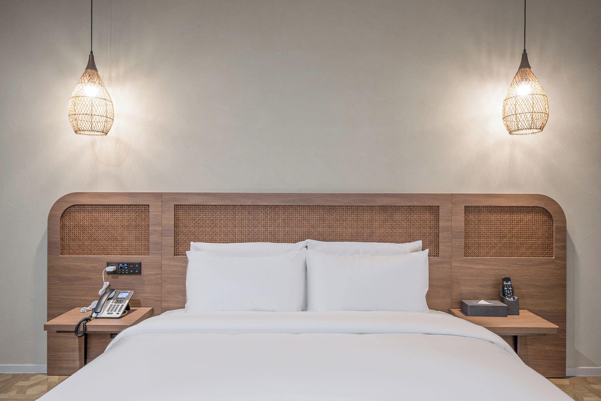 Una habitación de hotel con cabecera de madera, cama con ropa de cama blanca, mesita de noche con teléfono y dos lámparas colgantes.